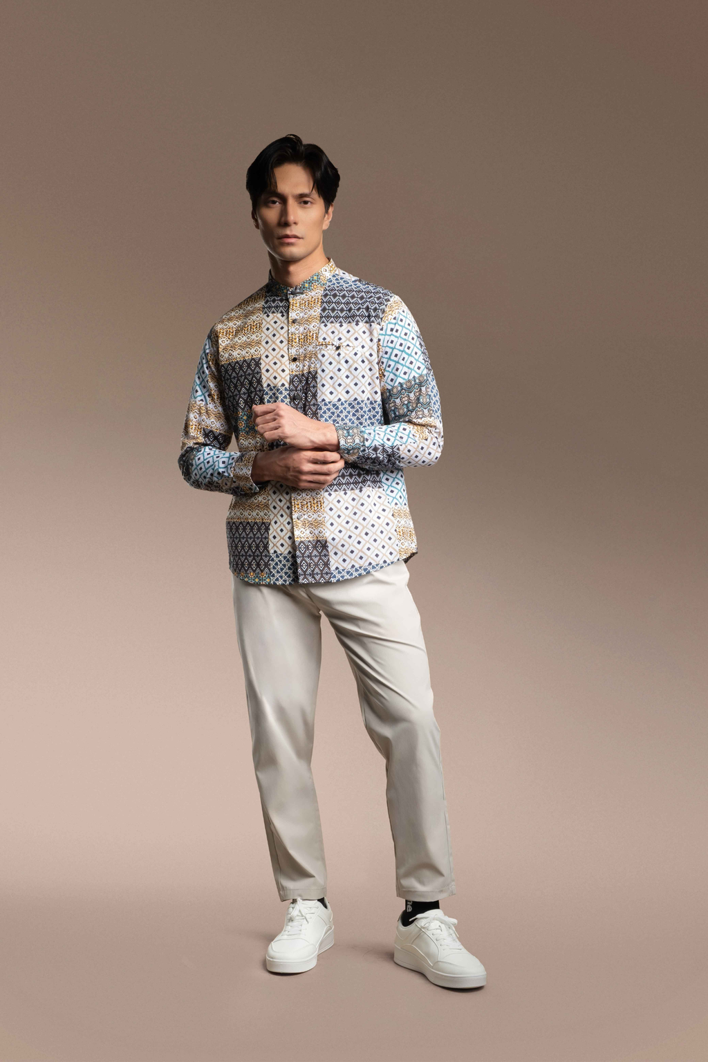 A man modeling a Onésimus Men's Casual Shirts: Geometric Prints paired with light beige trousers and white sneakers, against a solid warm brown studio background.