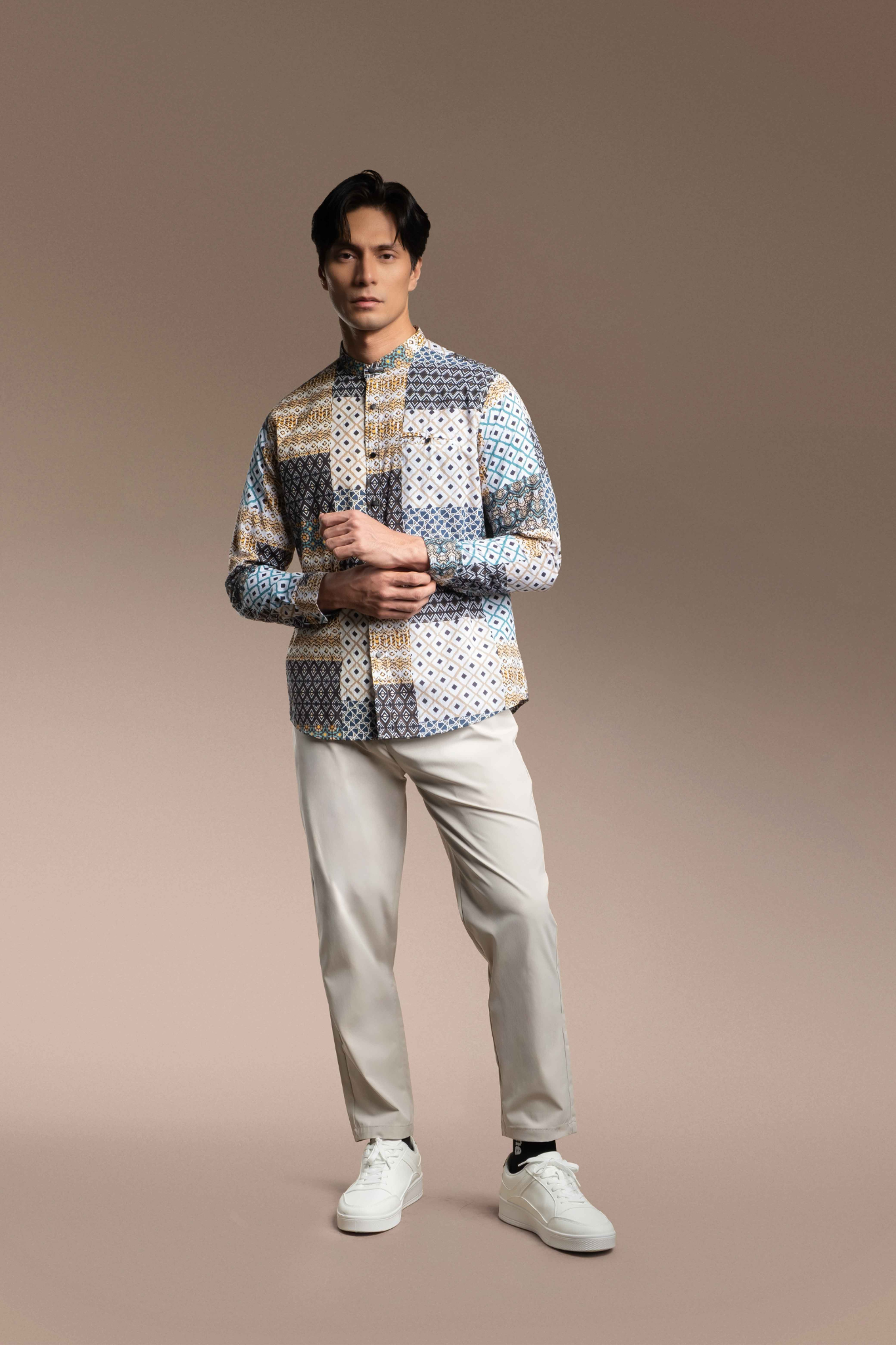 A man modeling a Onésimus Men's Casual Shirts: Geometric Prints paired with light beige trousers and white sneakers, against a solid warm brown studio background.